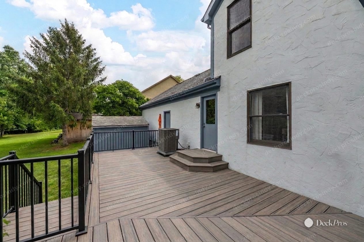 The same wrap-around deck after: warm grey Trex composite decking laid with a mitred border, black aluminum railing, and a rebuilt step up to the side door.