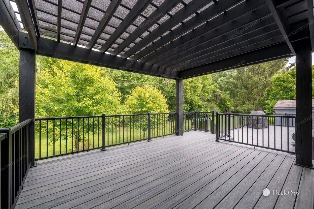 The same pergola deck after: grey Trex composite decking, black aluminum railing with slim balusters, and the pergola restained charcoal, open to the green treeline beyond.